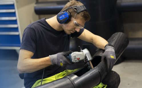 En konsentrert produksjonsarbeider med vernebriller og hørselvern sitter lent over et rør med et måleredskap i høyre hånd.