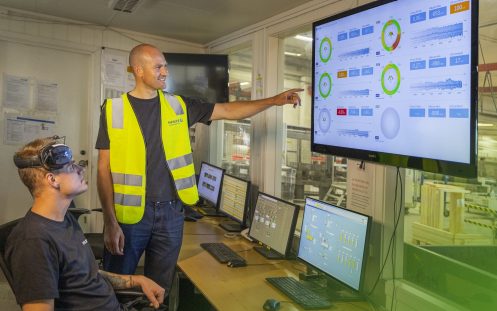 En person står oppreist og peker på en skjerm, mens en annen sitter i en stol mens de betrakter skjermen.