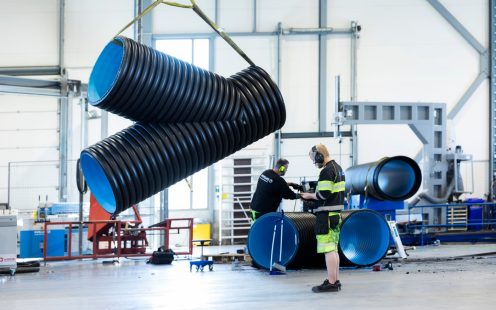 Et stort Y-formet rør hengende i luften etter en gul stropp, med en produksjonsansatt i arbeidsklær ved siden av.