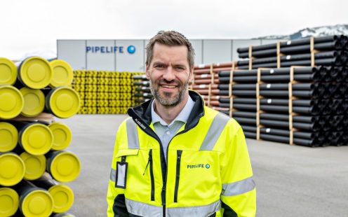 En smilende person i refleksjakke med stablede rør i bakgrunnen.