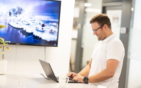 En smilende administrasjonsarbeider ikledd hvit kortermet skjorte sitter opptatt foran en laptop.