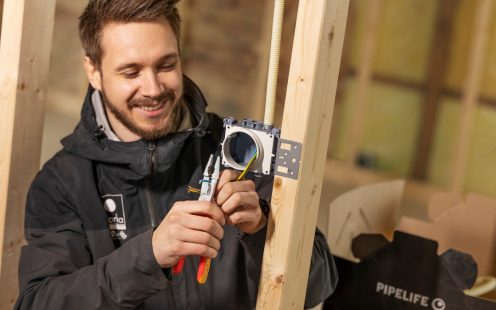 Smilende person lent mot en innendørs bjelke med eksponert elektronikk, med en tang i den ene hånda og ledninger i den andre.