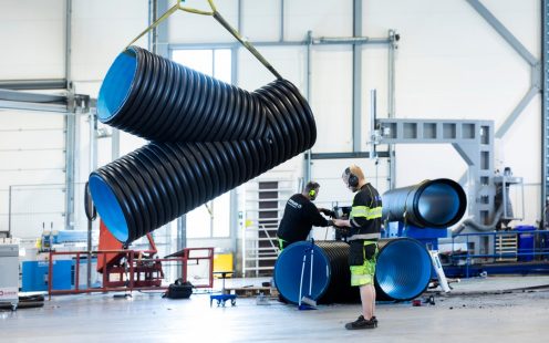 Et stort Y-formet rør hengende i luften etter en gul stropp, med en produksjonsansatt i arbeidsklær ved siden av.