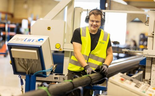 Smilende produksjonsarbeider med refleksvest og hørselvern som har hendene lenende på et sort plastrør.