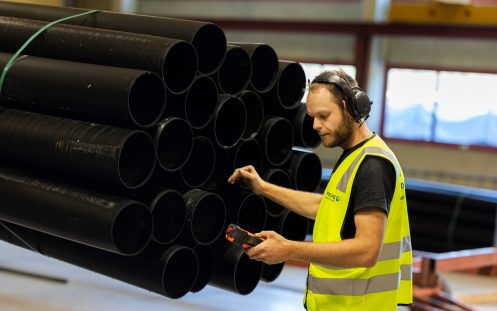 Konsentrert produksjonsarbeider med refleksvest og hørselvern som har én hånd lenende på en hengende bunt med sorte plastrør.