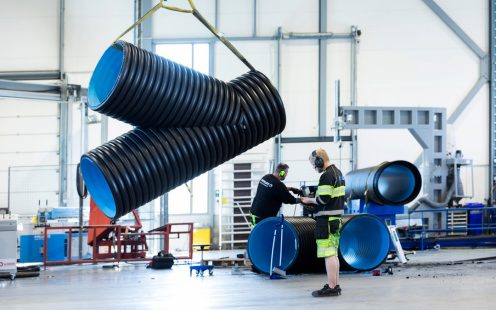 Et stort Y-formet rør hengende i luften etter en gul stropp, med en produksjonsansatt i arbeidsklær ved siden av.