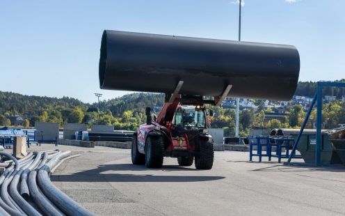 En teleskoplaster kjørende gjennom en anleggsplass med et stort sort rør i gaflene.