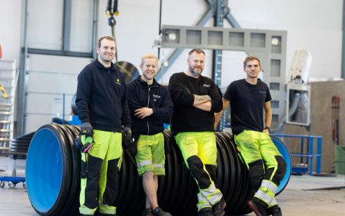 Fire produksjonsansatte, alle ikledd sort overdel og gule refleksbukser, ser smilende inn i kamera mens de lener seg på et stort sort rør.