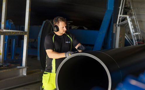 En fokusert produksjonsansatt ikledd sort t-skjorte, hansker og hørselvern måler utsiden av et stort sort rør.