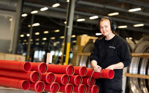 En produksjonsansatt ikledd sort genser og hørselvern ser smilende inn i kamera, med hendene lenende på en rekke røde rør.