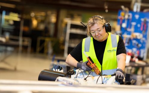 En fokusert produksjonsansatt ikledd sort t-skjorte, gul refleksvest og hørselvern står framoverlent over et rør med et verktøy i den ene hånden.