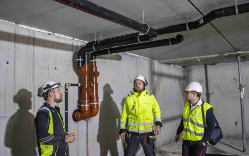 Tre arbeidere i refleksjakker står inne i et rom og ser opp på et rørsystem som løper langs taket.