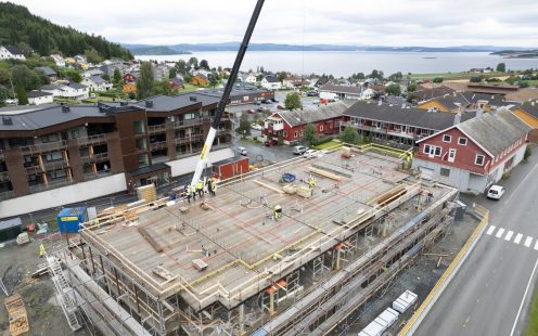 Oversiktsbilde av anleggsarbeid som foregår på taket til et bygg, med en gate i forgrunnen og en fjord i bakgrunnen.