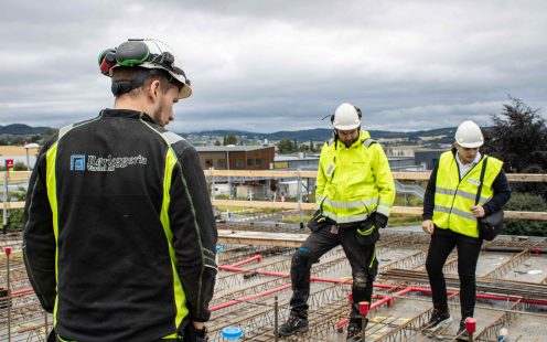 Tre arbeidere i refleksjakker står ute på et anleggsområde og ser ned på arbeidet som er gjort i bakken.
